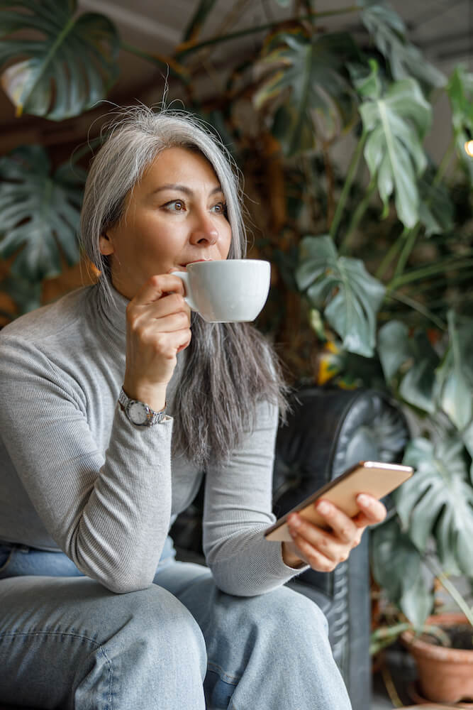 Woman sipping coffee while looking away thoughtfully, holding a smartphone in a plant-filled room.
